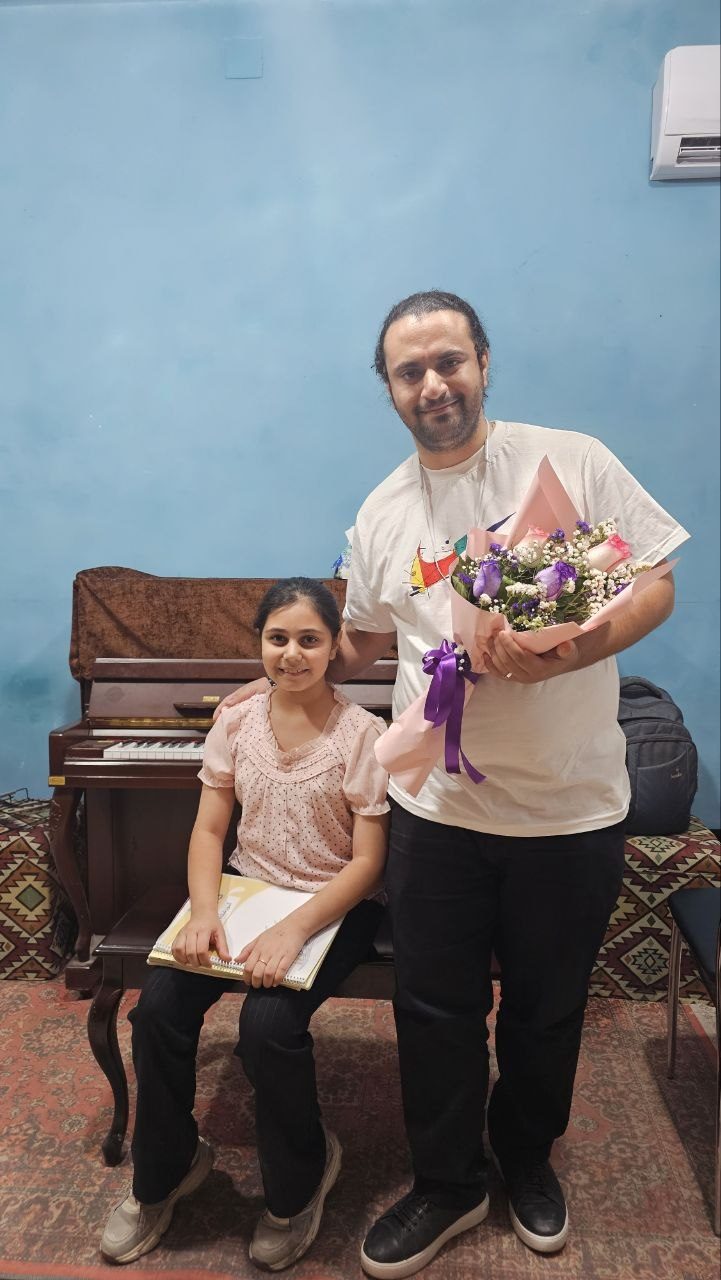 A young student seated at the piano with Mohammadali Rezaei holding flowers